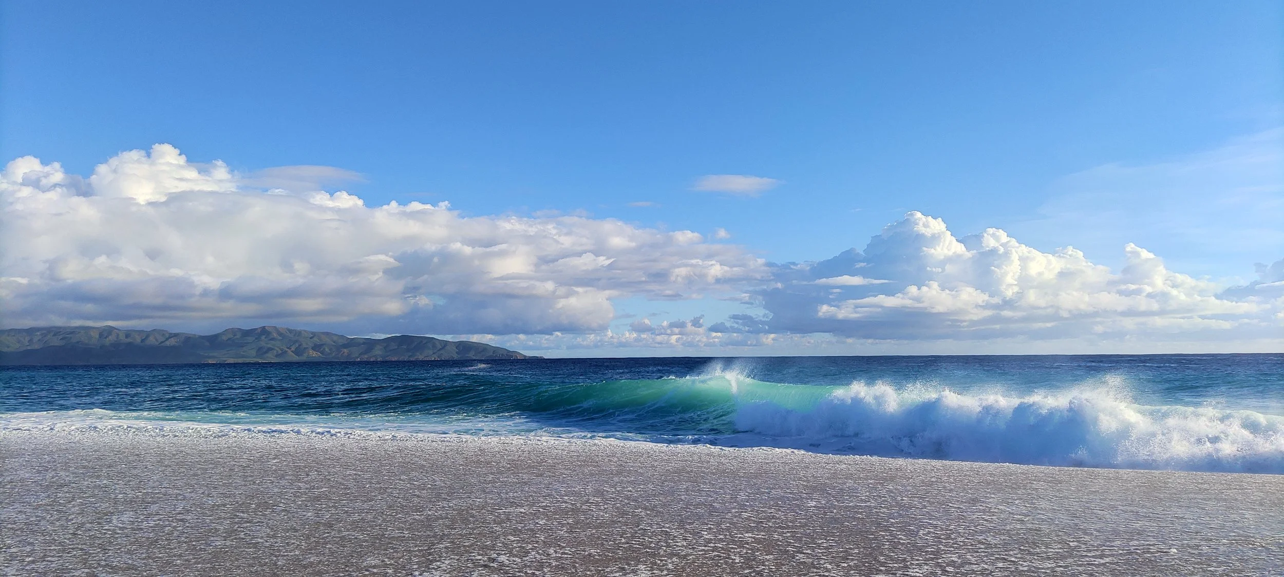 A beach scene with waves crashing on the shore, a distant Cerralvo Island mountain range, blue sky with scattered clouds.