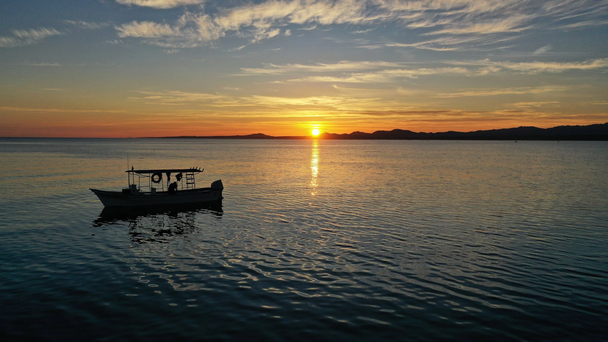 A small boat floating on calm water during sunset, with mountains in the background and a colorful sky with clouds.