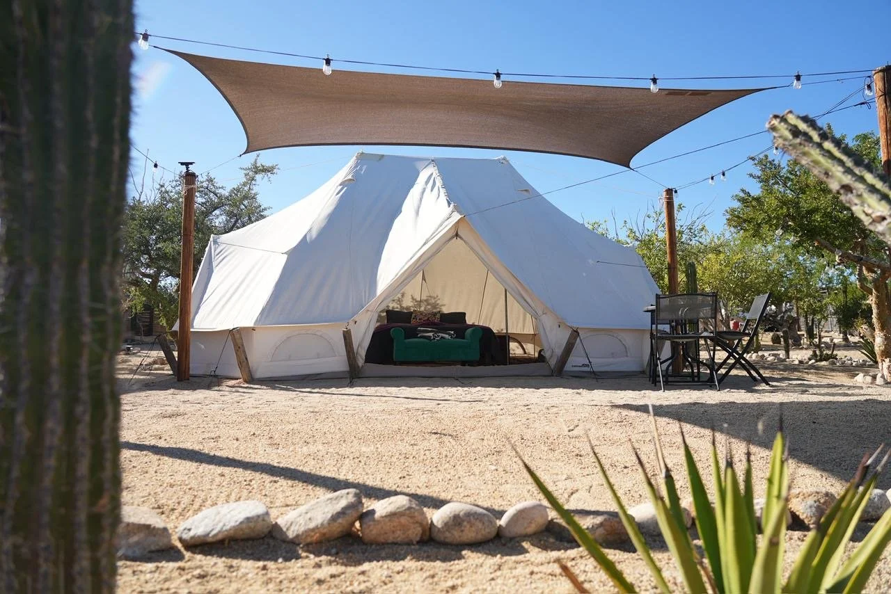 A white canvas tent set up in a desert area with a gravel ground, surrounded by trees and plants. There is an outdoor seating area with a table and chairs, and a shaded canopy overhead with string lights. The sky is clear and blue.