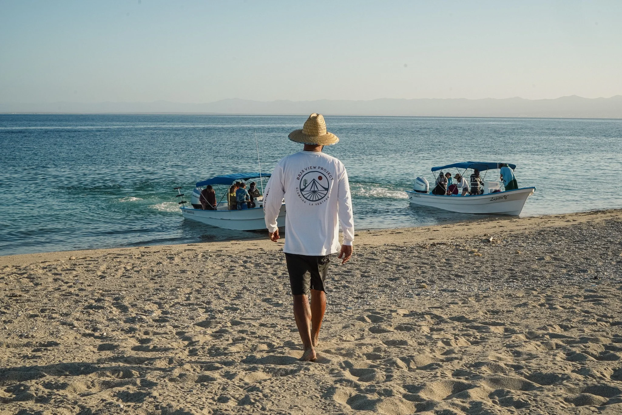 A person wearing a wide-brimmed straw hat and a white shirt with a camping logo walks along a sandy beach towards two boats with people on board. The water is calm and the sky is clear.