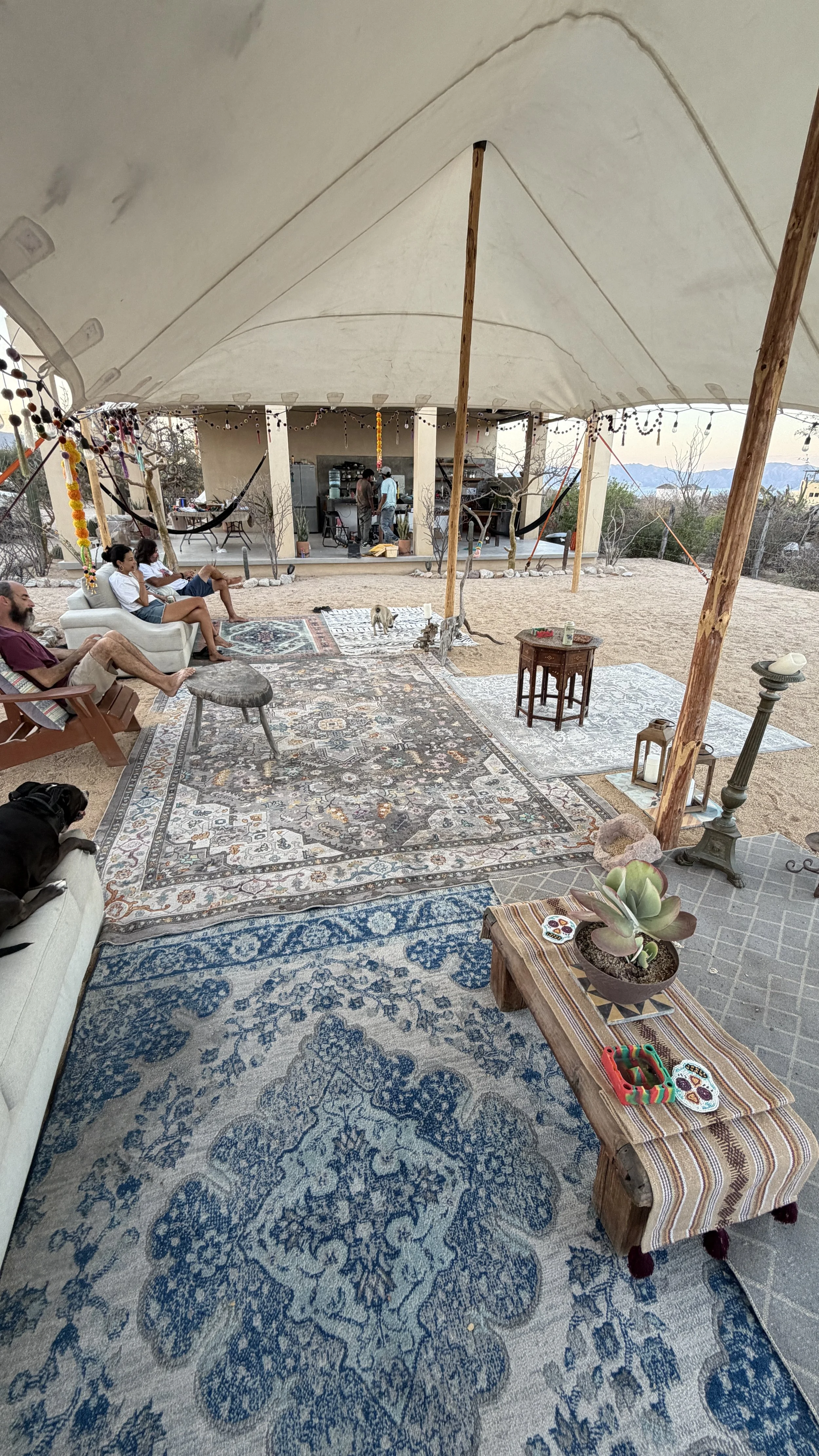 Outdoor gathering under a large white canopy with people sitting on sofas and chairs, patterned rugs on the ground, and a small table with potted plants and decorative items; in the background, there is a house with a patio and mountain view.