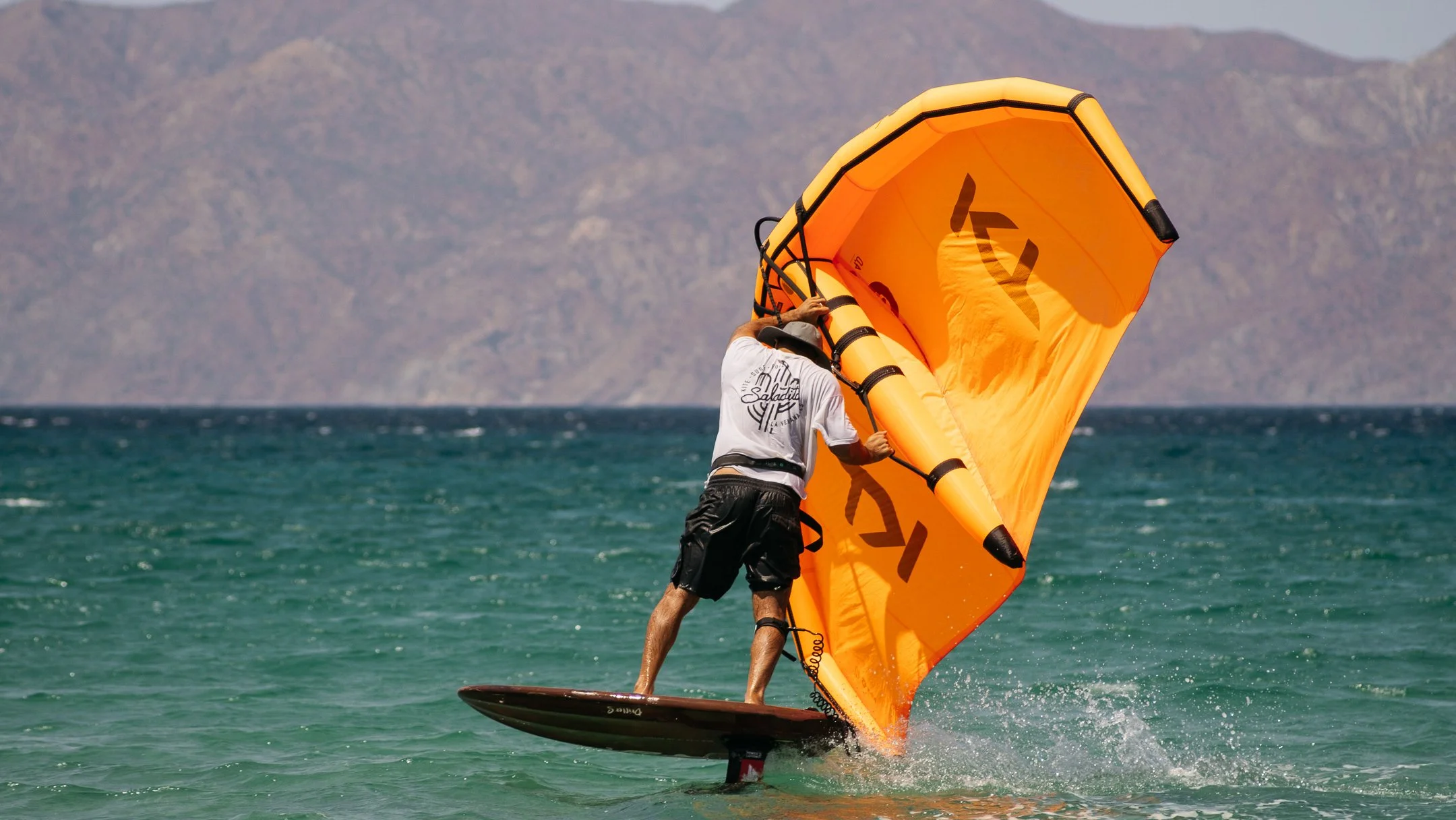 Saladita kite school & the foil club founder Ando Flores  engaged in wingfoiling on the ocean, holding an orange KT sail, with a backdrop of cerralvo island at La Ventana.