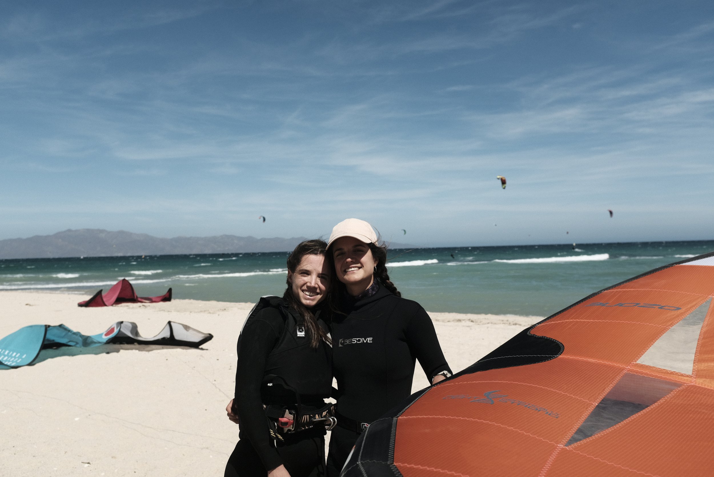 Two women smiling on a beach with Wingfoil and kiteboarding equipment, blue sky, and ocean with kite surfers in the background just abouth to start their lessons with Saladita Kite School.