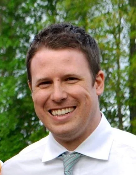 Young man smiling outdoors with green trees in the background, wearing a white shirt and a light-colored tie.