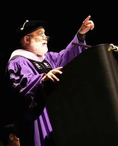 Jack Dillenberg in academic regalia standing at a podium, gesturing with his right hand while speaking.
