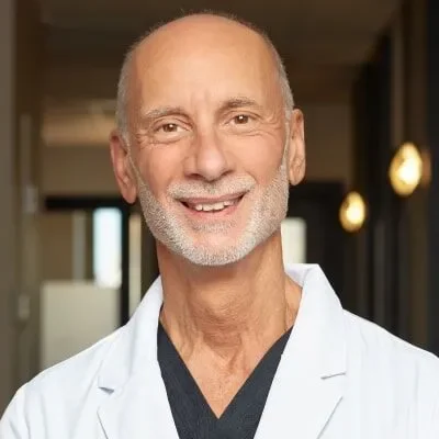 A smiling older man with a bald head and gray beard, wearing a white lab coat, in an indoor setting.