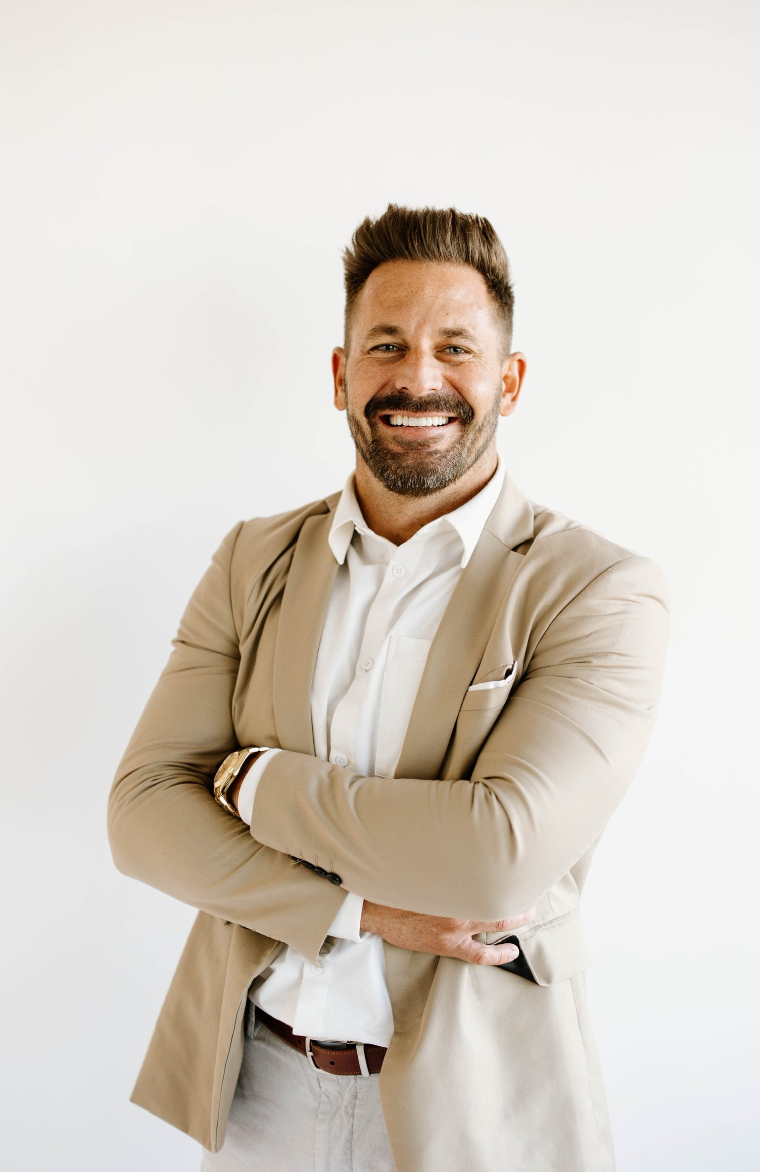 A man with a beard, dressed in a beige blazer and white shirt, smiling with arms crossed against a plain white background.