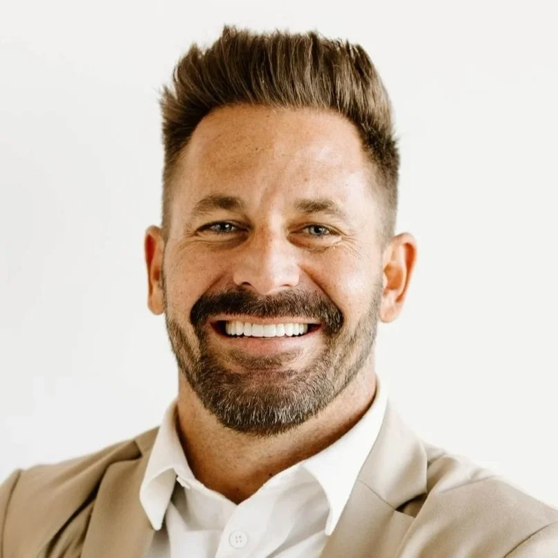 A smiling man with a beard and short brown hair, wearing a beige blazer and white shirt, against a plain white background.