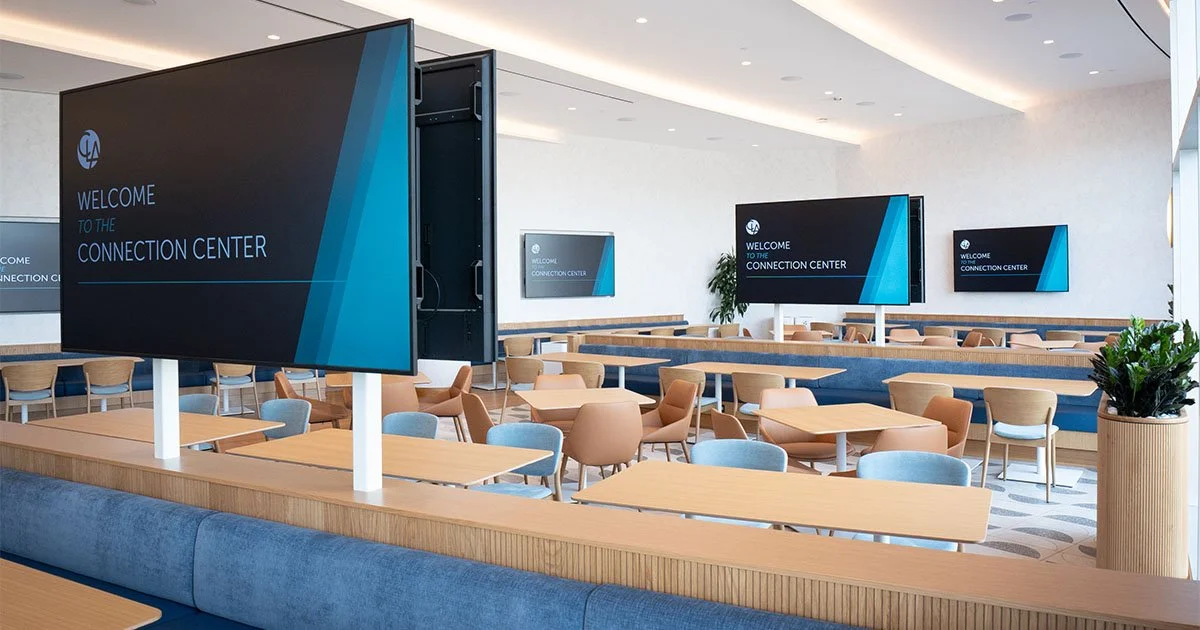Modern conference or meeting room with multiple large screens displaying 'Welcome to the Connection Center', round and rectangular tables, and chairs. The space is bright with natural light and has potted plants for decor.