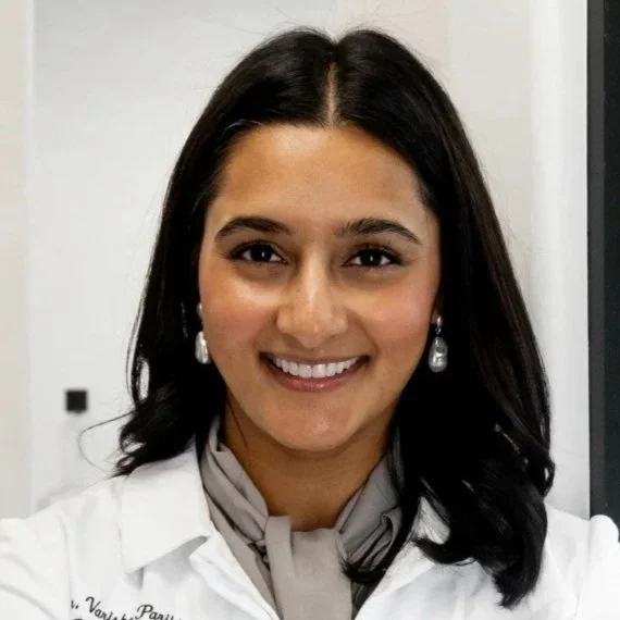 A woman with dark hair and earrings smiling at the camera, wearing a white coat and a light scarf.