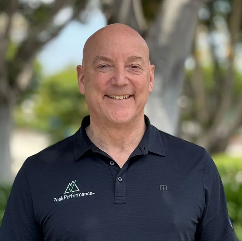 A smiling bald man wearing a navy polo shirt with Peak Performance logo, standing outdoors with blurred trees in background.