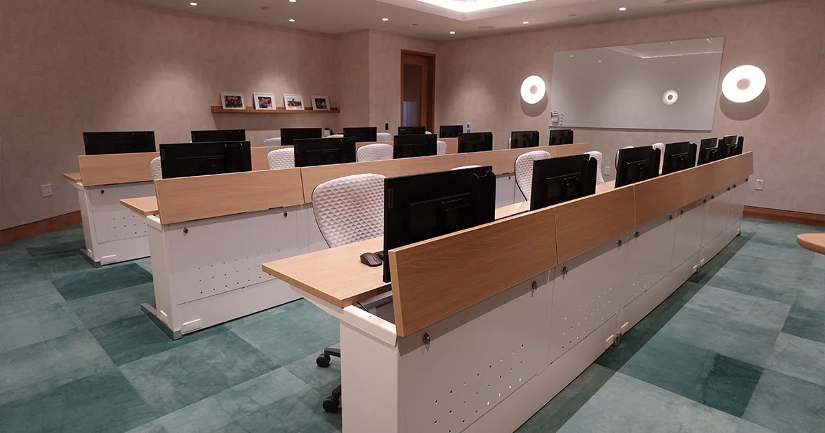 Empty classroom with individual desks equipped with computers and chairs, a large mirror on the wall, and framed photographs on a shelf.