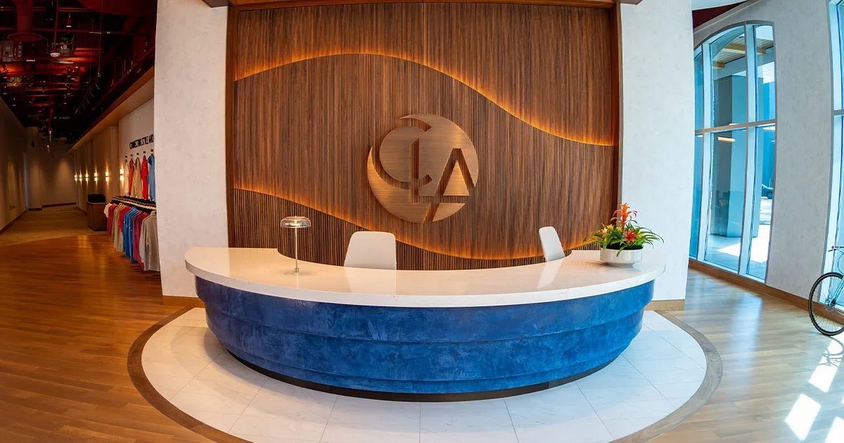 Hotel reception desk with a white marble top, blue velvet base, and a modern wooden wall with an illuminated logo behind it, situated in a bright lobby with wood flooring, large glass windows, and a clothing display to the left.