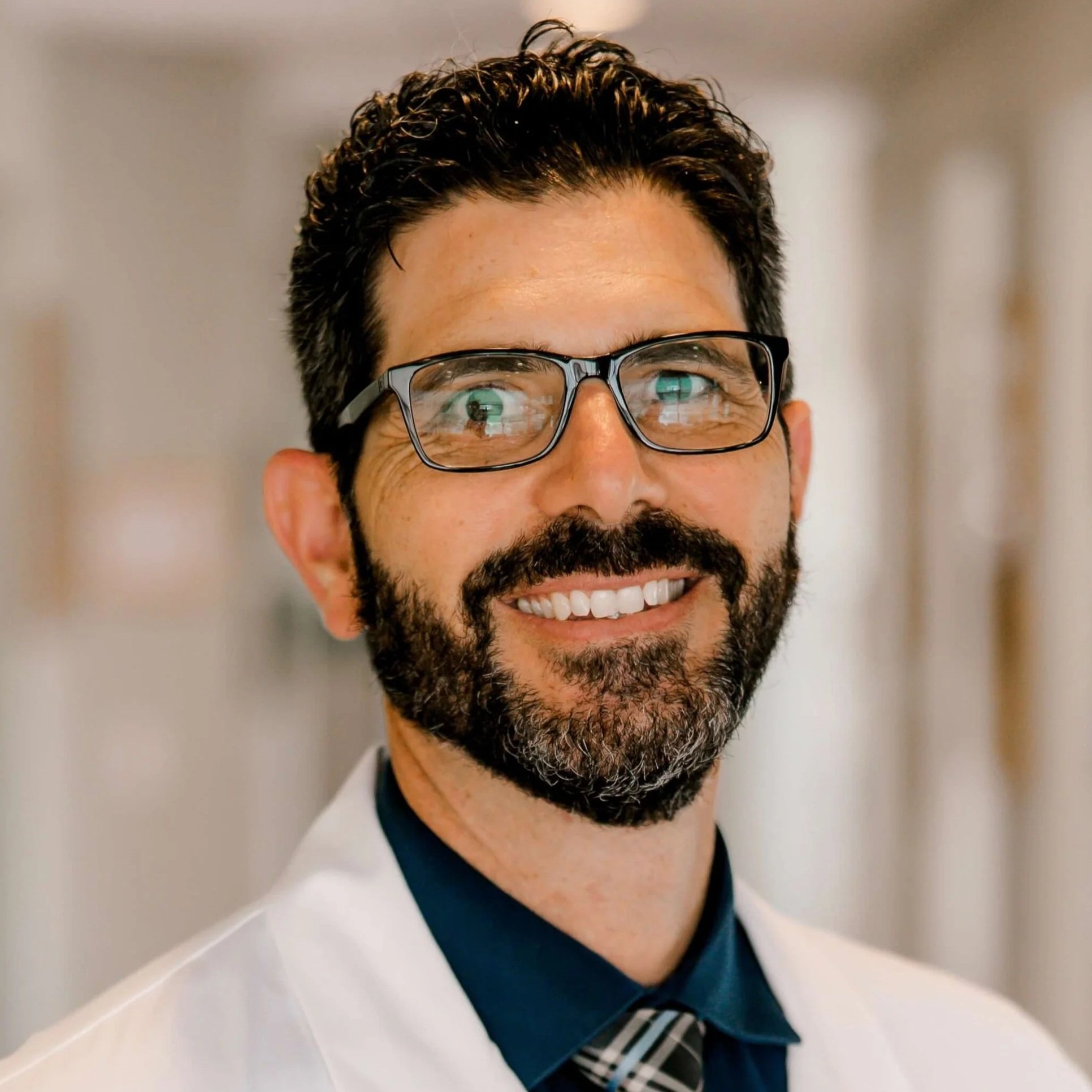 Close-up of man with dark curly hair, glasses, and a beard, smiling in a professional setting.
