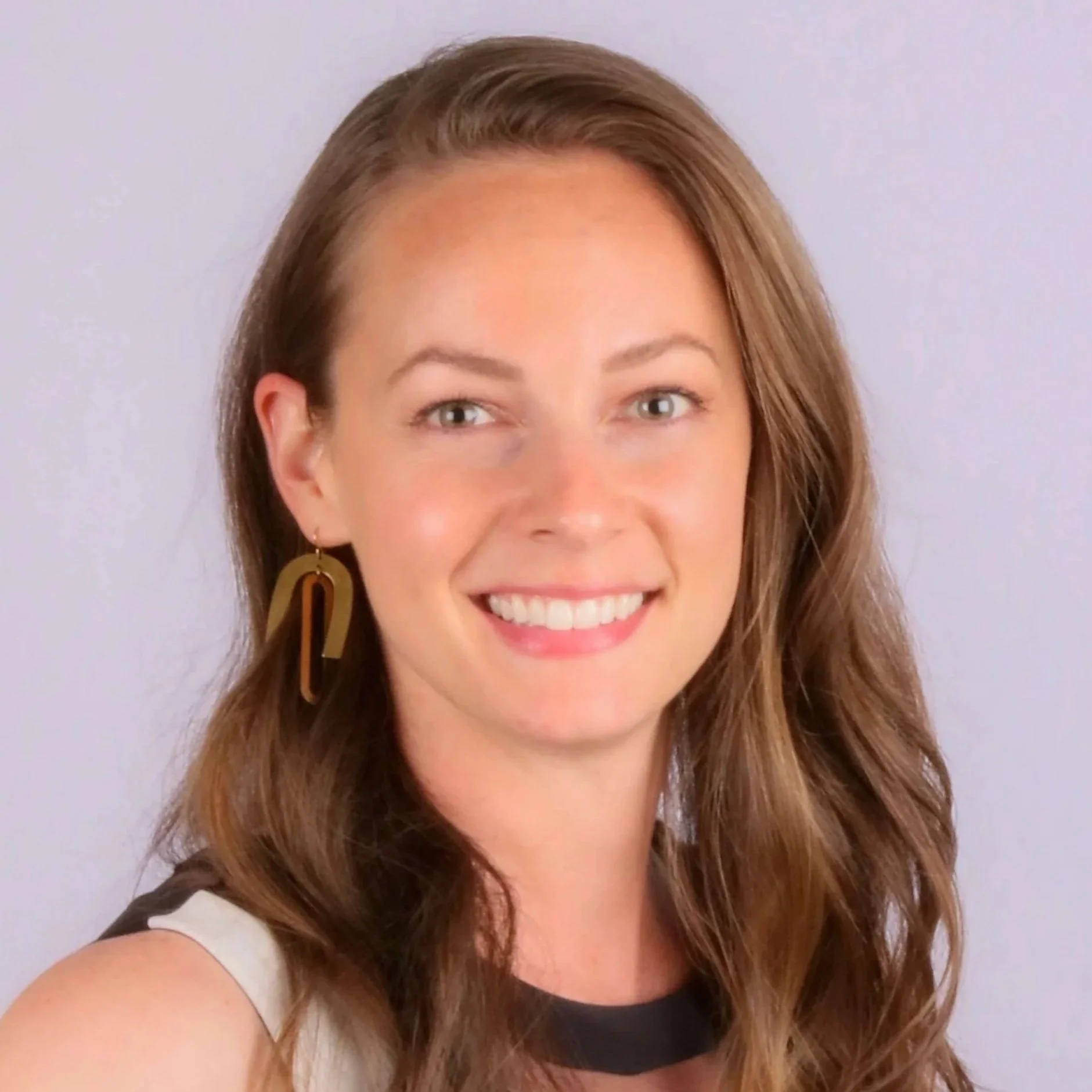 Close-up of a woman with light brown, wavy hair, smiling, with gold earrings, wearing a black top with a beige or cream-colored sleeve, against a plain light background.