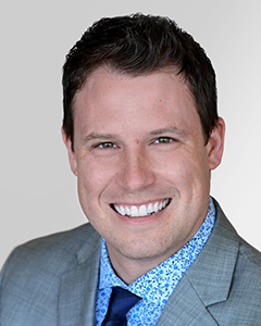 Professional headshot of a man with short dark hair, smiling, wearing a grey suit jacket, light blue shirt, and dark blue tie, against a plain grey background.
