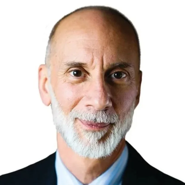 Close-up portrait of an older man with a bald head, gray beard, and blue eyes, wearing a dark suit jacket and light blue shirt, smiling slightly against a white background.