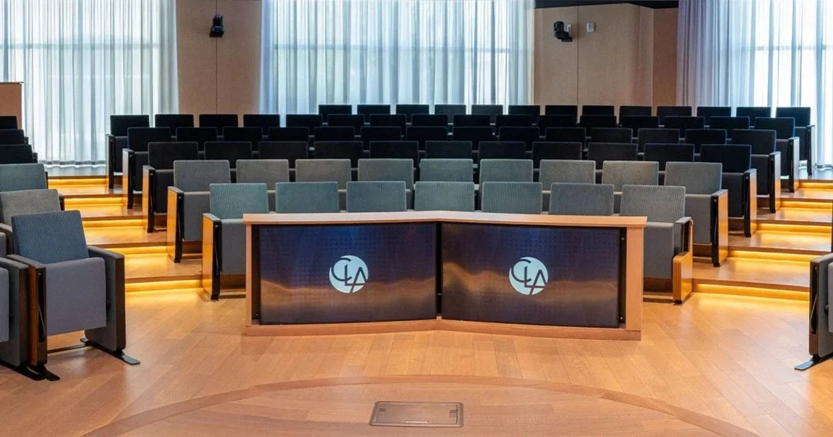 Empty conference room with rows of chairs behind a desk with two digital screens displaying a logo.