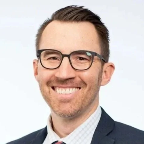 Headshot of a man wearing glasses, a suit, and a tie, smiling against a plain background.
