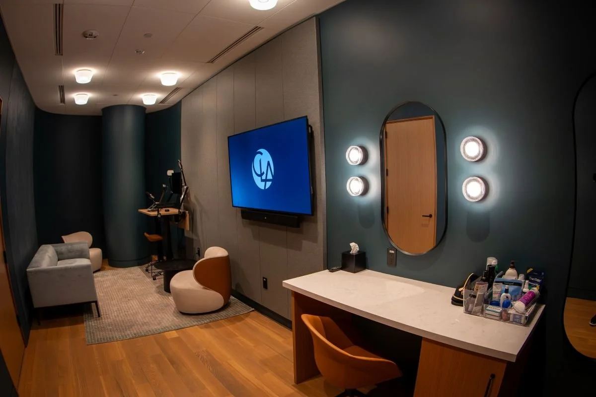 A modern room with a mounted flat-screen TV, a vanity with a mirror and wall lights, a small seating area with a sofa and two chairs, and a desktop with various personal care items.