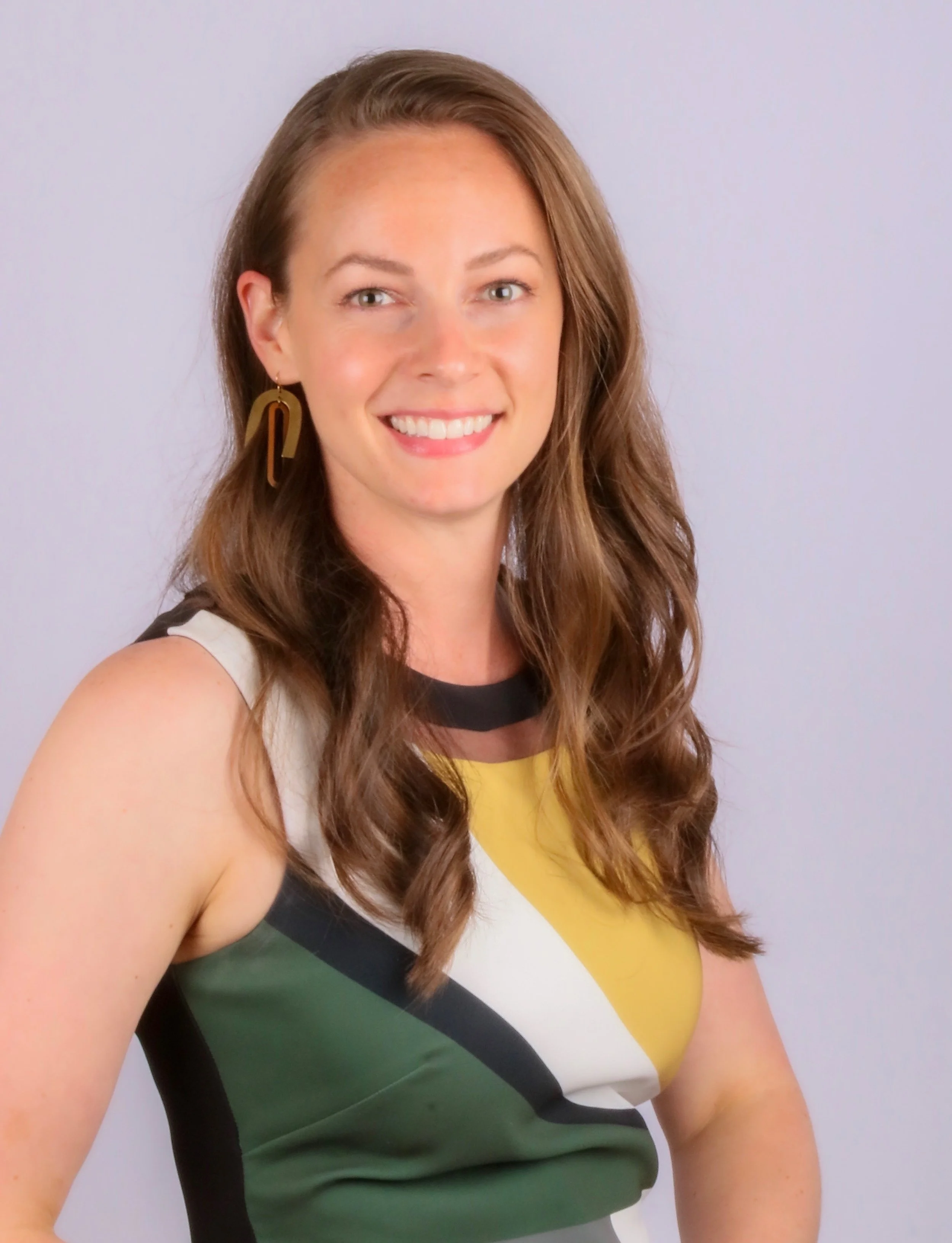 Close-up of a smiling woman with long, wavy brown hair, wearing a sleeveless top with black, yellow, white, and green geometric shapes, and gold earrings, against a light purple background.