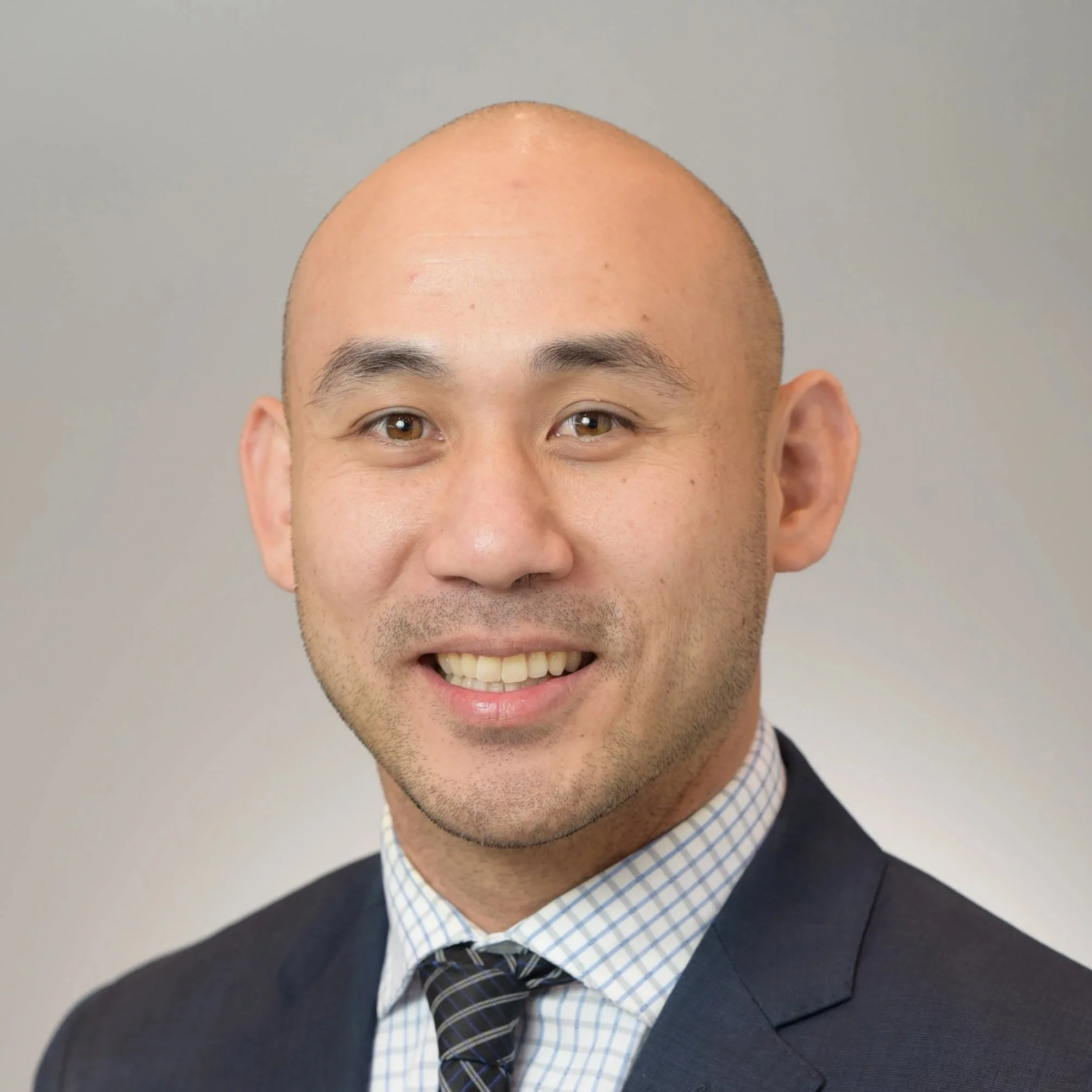 Close-up of a smiling man with a bald head, wearing a dark blazer, patterned shirt, and a tie, against a plain gray background.