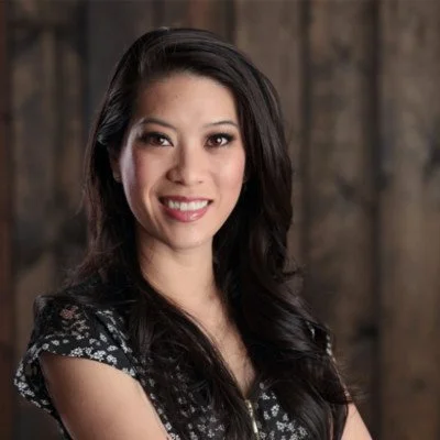 A woman with long dark hair smiling at the camera, wearing a black floral top, against a rustic wooden background.