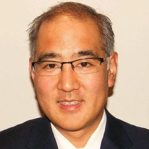 Close-up of a middle-aged man with short gray hair, wearing glasses, a dark suit, and a white shirt, against a plain white background.
