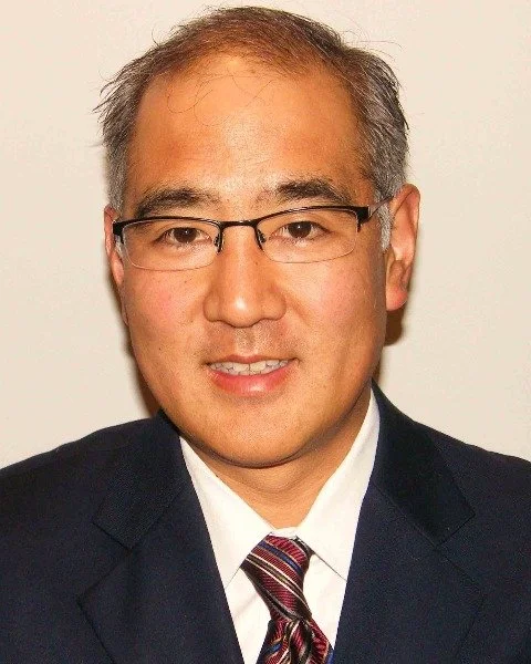 Portrait of a man wearing glasses, a dark suit, white shirt, and a striped tie, smiling against a plain background.