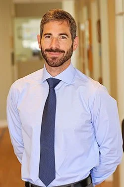 A man with a beard wearing a light blue dress shirt and dark tie, standing in a hallway with bookshelves and windows in the background.