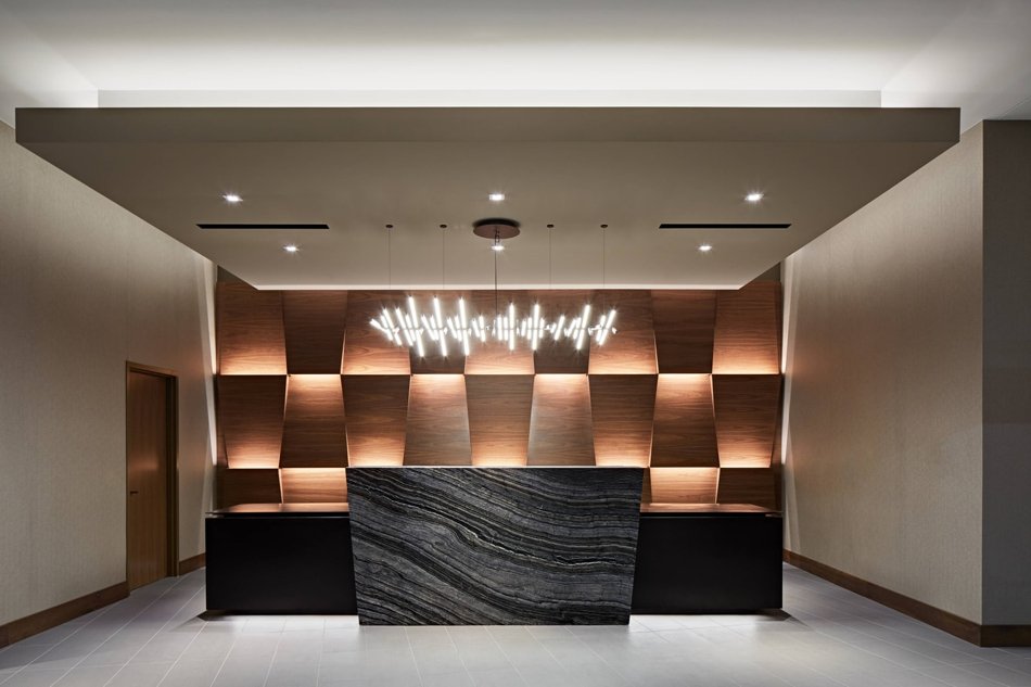 Modern hotel or office reception area with a marble front desk, geometric wooden wall panels, and a contemporary chandelier.