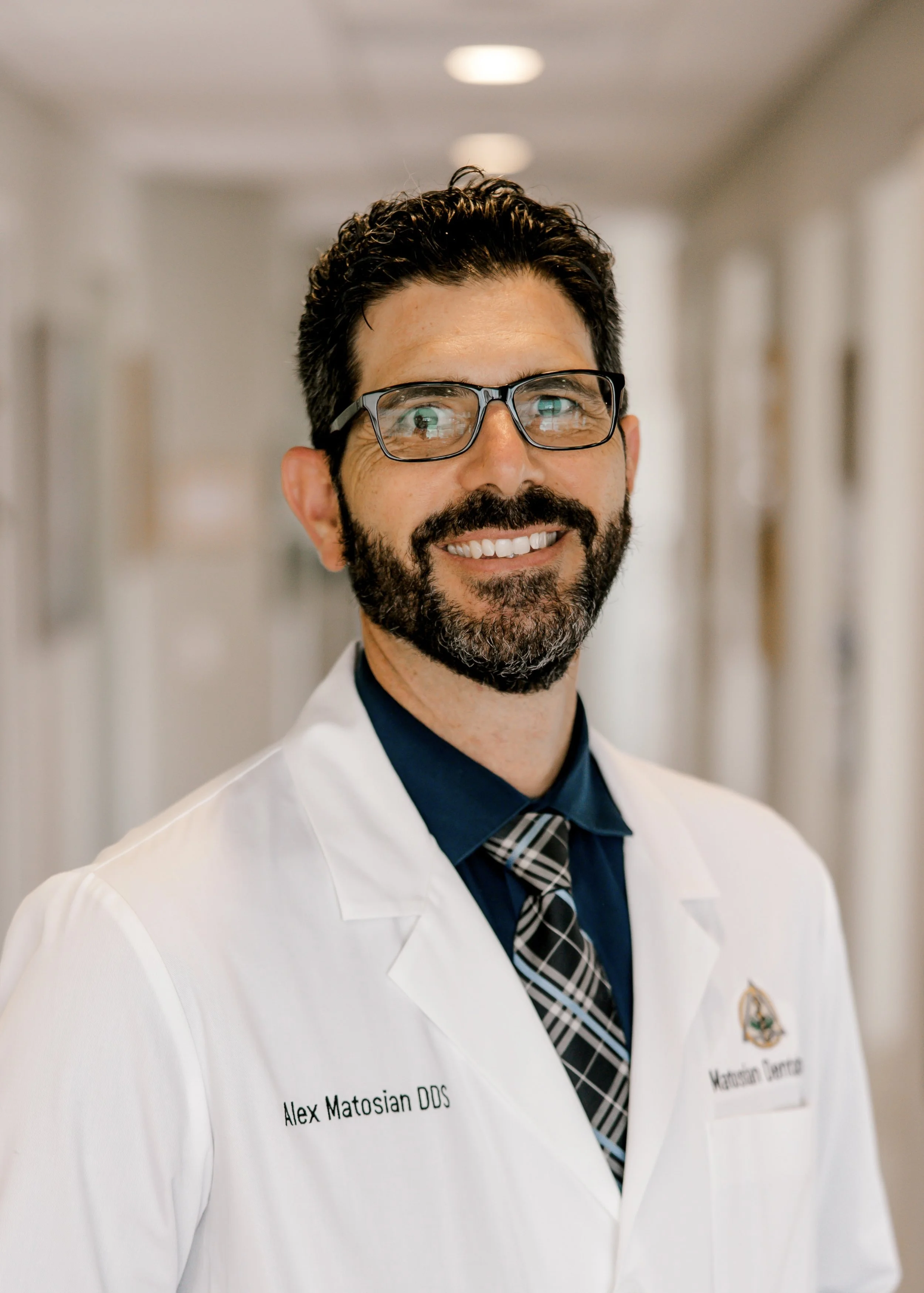 A man with dark hair, glasses, and a beard smiling in a hallway, wearing a white lab coat with a name tag that reads Alex Matosian DOS and another name tag with a logo.