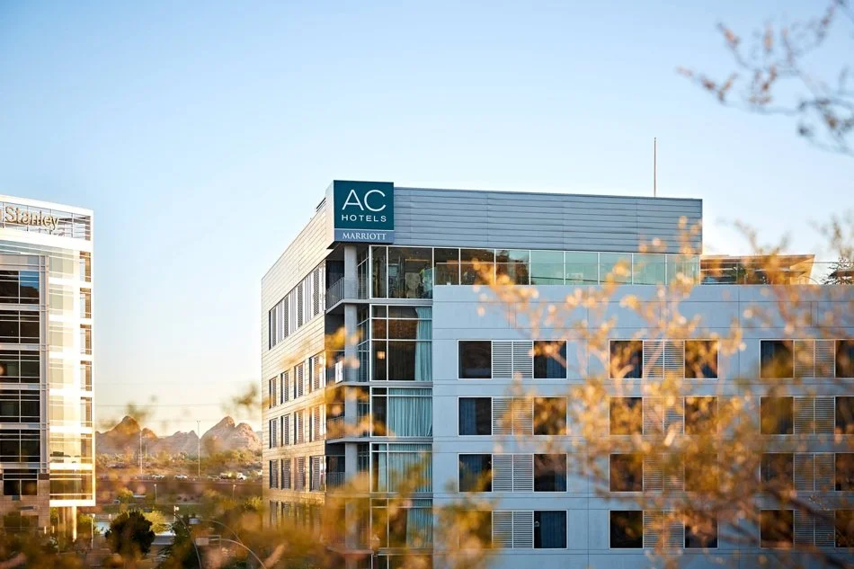 Modern hotel building with AC Hotels and Marriott signage, surrounded by nearby buildings and autumn trees, under a clear blue sky.