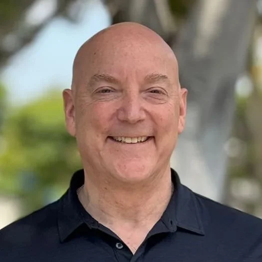 A smiling bald man in a black collared shirt standing outdoors with trees in the background.