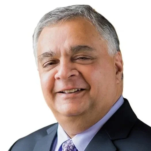 A middle-aged man with gray hair smiling, dressed in a dark suit, light blue shirt, and purple tie, against a white background.