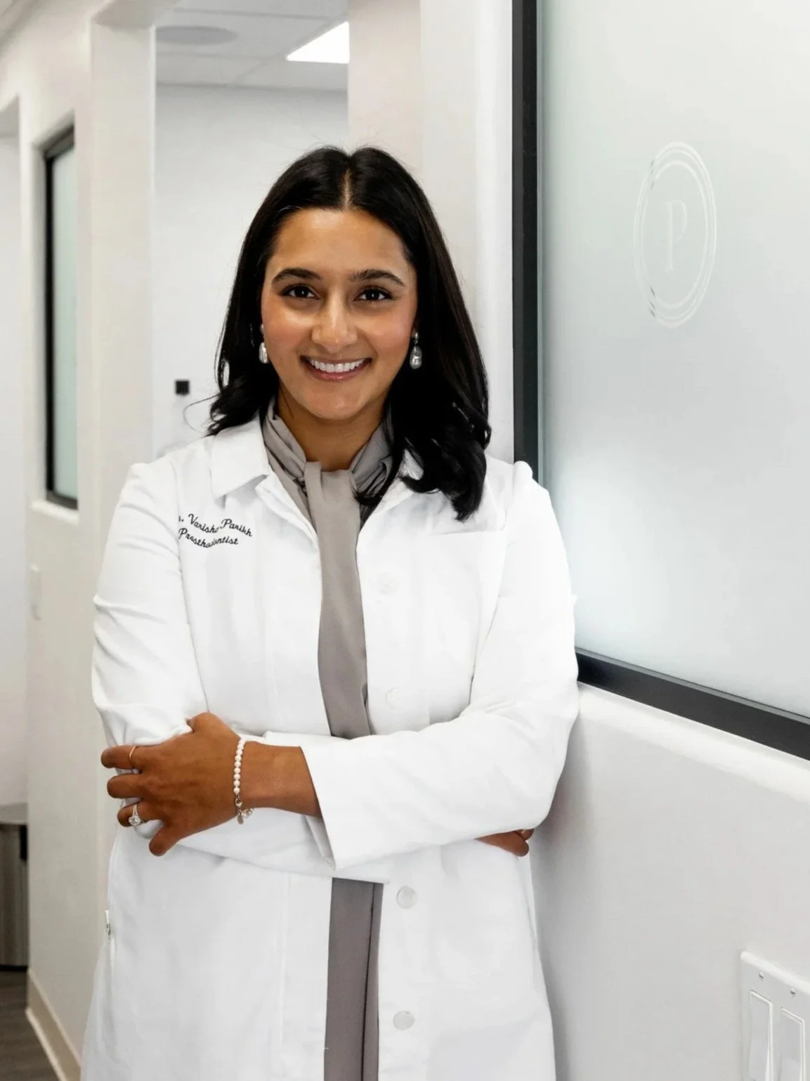 A woman in a white medical coat standing in a medical office, smiling with her arms crossed, in front of a whiteboard and a hallway.