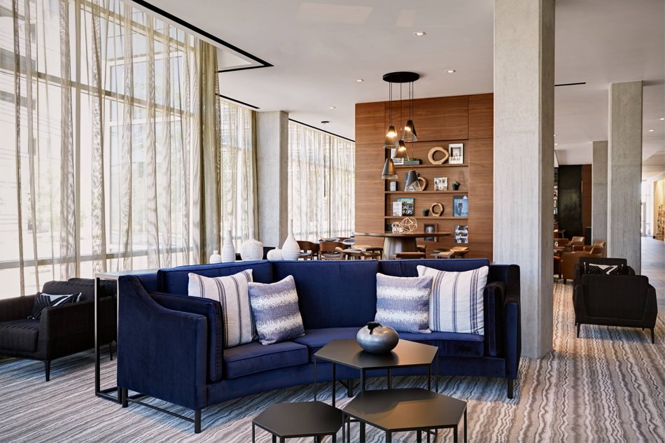 Modern hotel lobby with a navy blue sofa, decorative pillows, and a coffee table, surrounded by large windows with sheer curtains and stylish shelving on an accent wall.