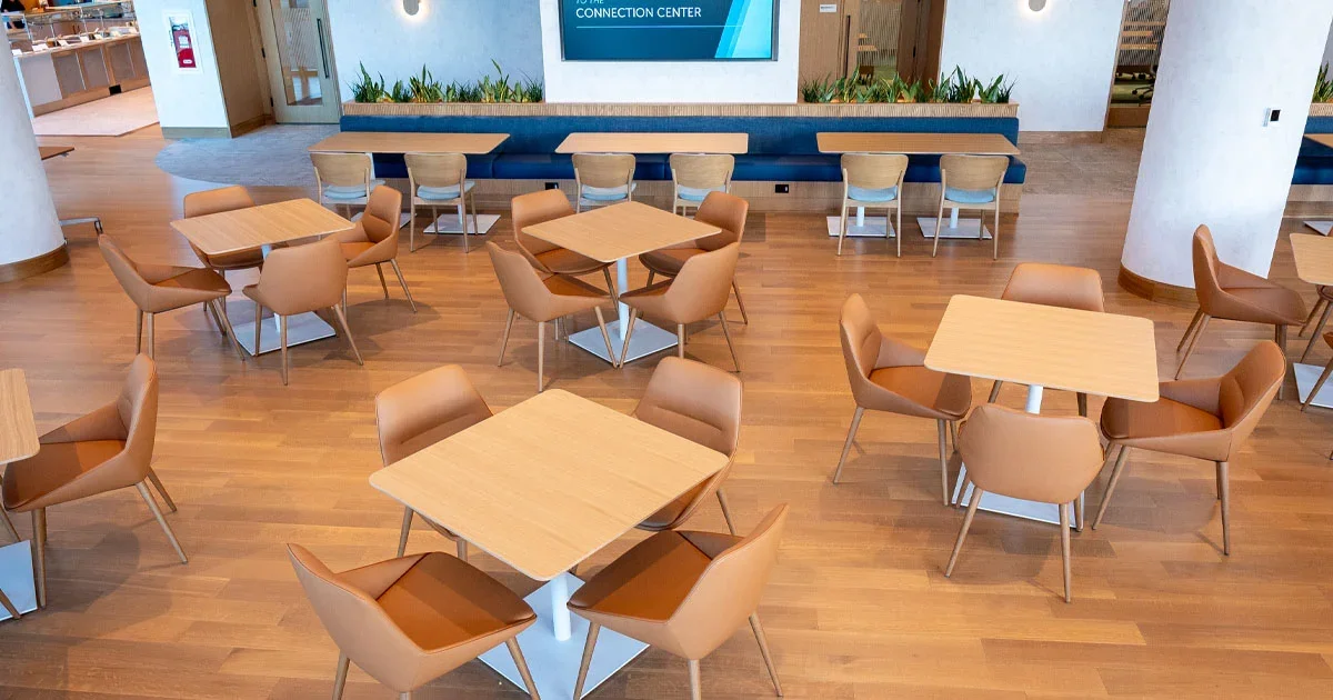 An empty modern lounge with wooden tables and tan chairs, a blue banquette along the back, plants, and a large screen on the wall.