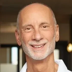Headshot of Kirk Pasquinelli, wearing a white shirt, standing indoors with windows in the background.