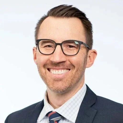A man with short brown hair, glasses, and a beard, smiling in a professional suit with a white dress shirt and striped tie.