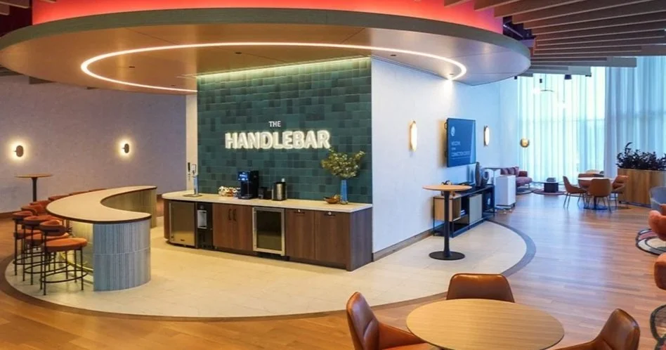 Modern lounge area with a curved bar counter, seating, and a sign reading 'The Handlebar', with a coffee station and a large window with sheer curtains.