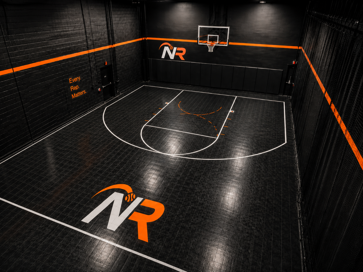 An indoor basketball court with black tiles, black walls, and orange and white markings. The court features the NR logo on the floor and on the wall, with a basketball hoop at one end. The slogan "Every. Rep. Matters." is displayed on the wall to the left.