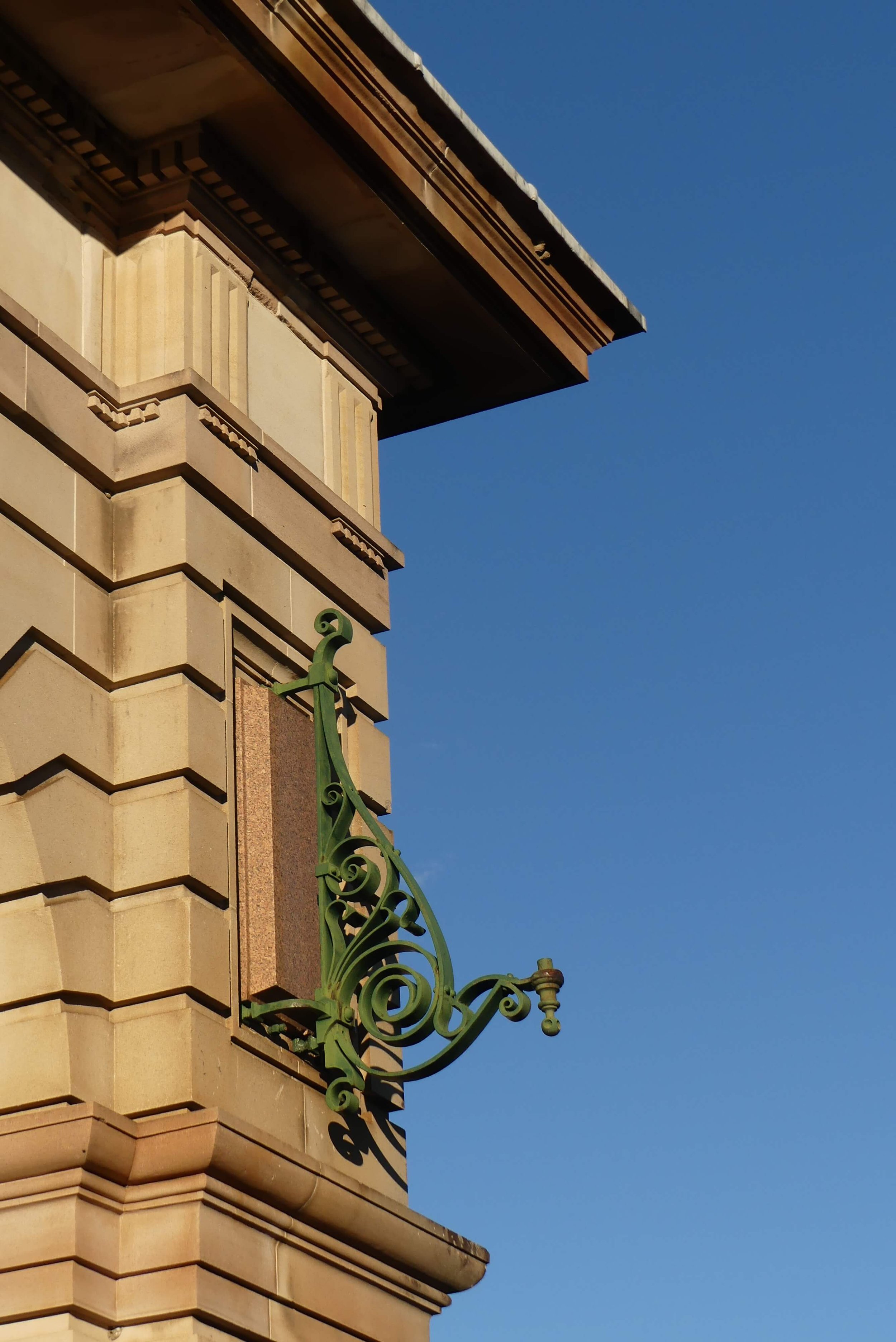 A corner of a building with ornate stonework and a decorative green wrought iron sign bracket against a clear blue sky.