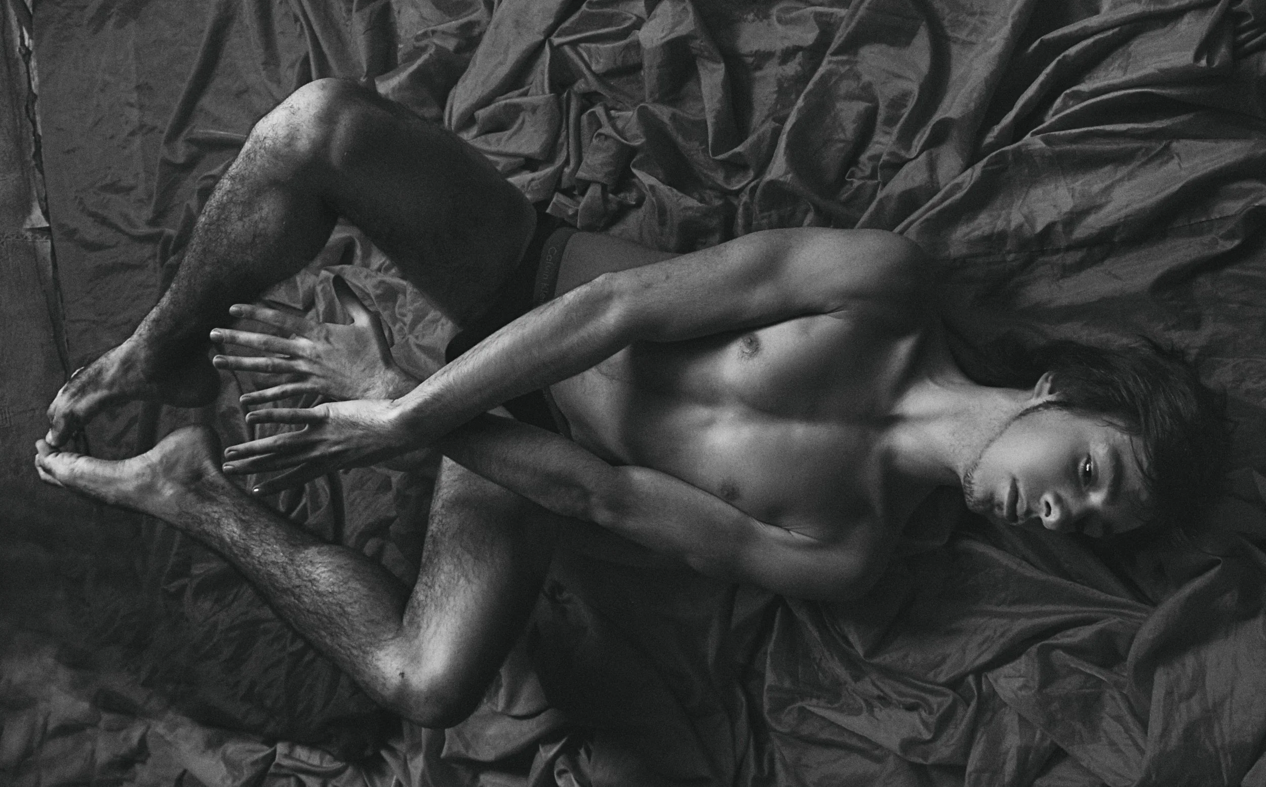 Black and white photograph of a shirtless man laying on a bed with black sheets. His legs are crossed, and his face is turned slightly to the side with his eyes open.