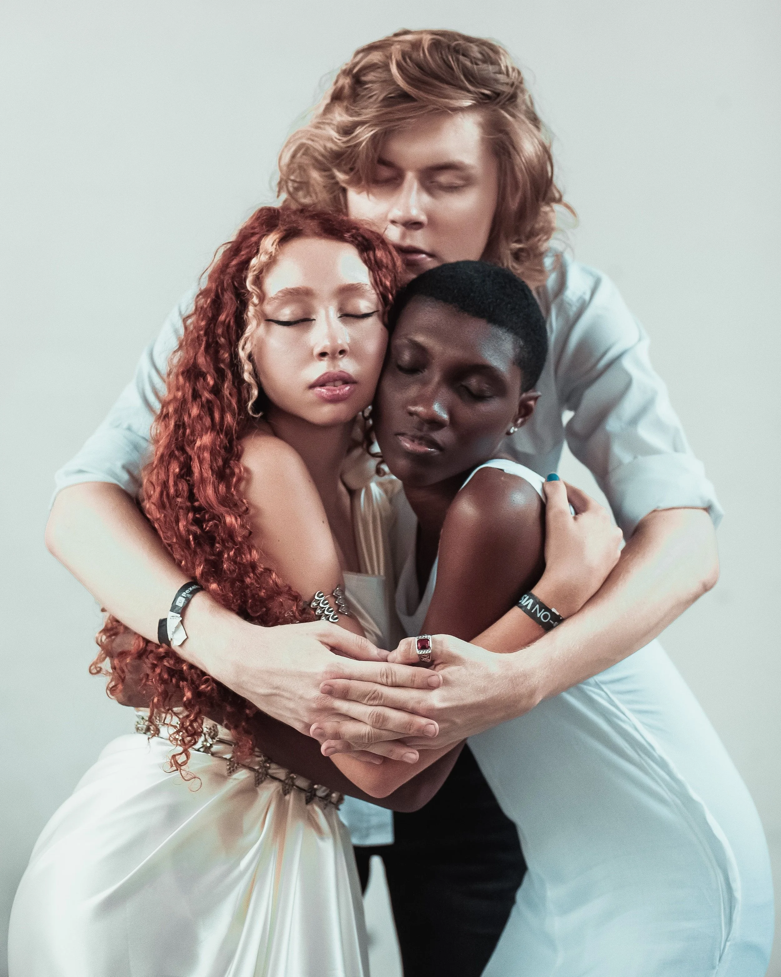 Four people, two women and two men, are hugging each other with their eyes closed, standing against a plain white background.