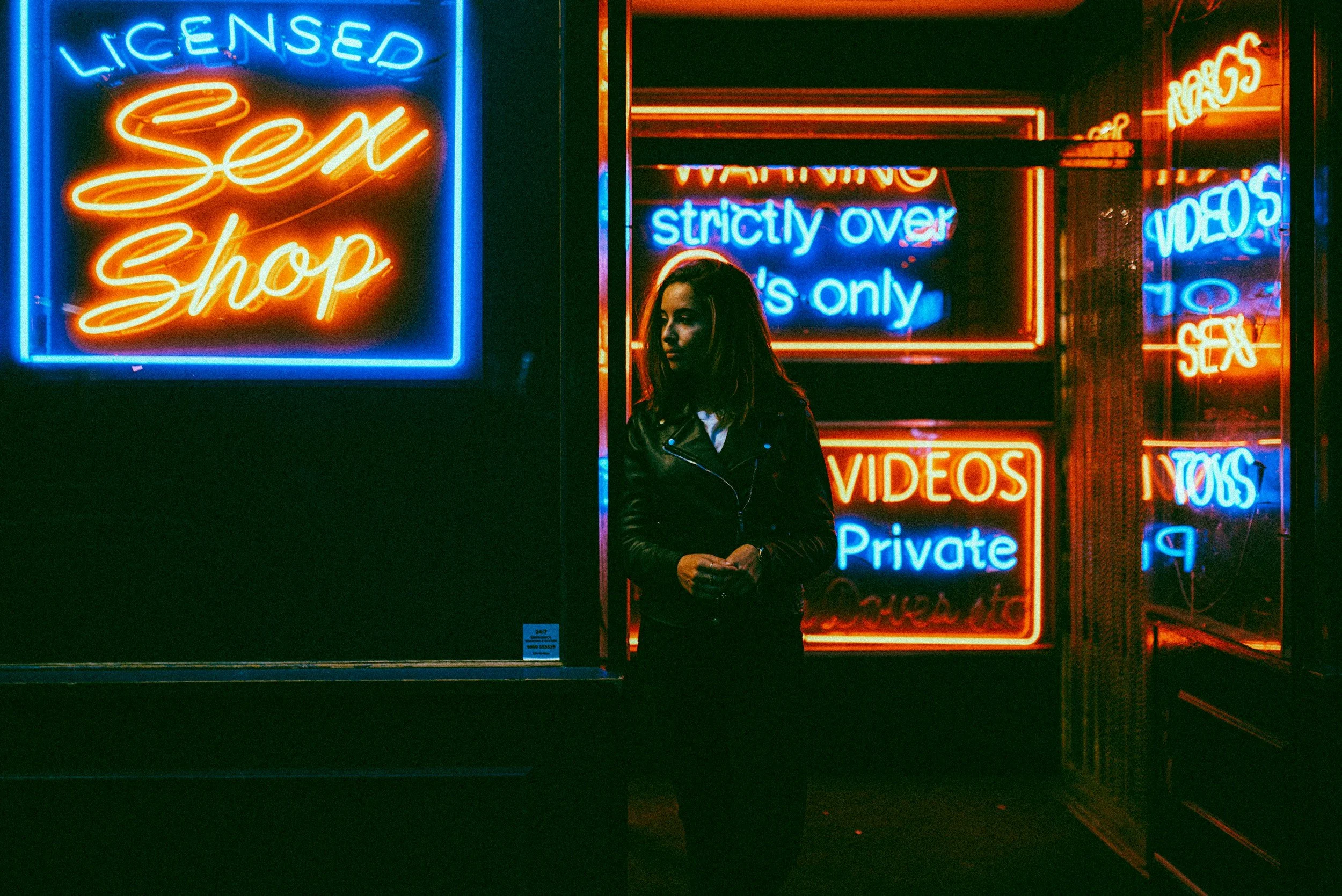 A woman with long hair is standing in front of neon signs that advertise an adult store. The signs include messages like 'LICENSED Sex Shop', 'strictly over 18s only', 'VIDEOS', and other adult entertainment content. The scene is dark, illuminated by the colorful neon lights.