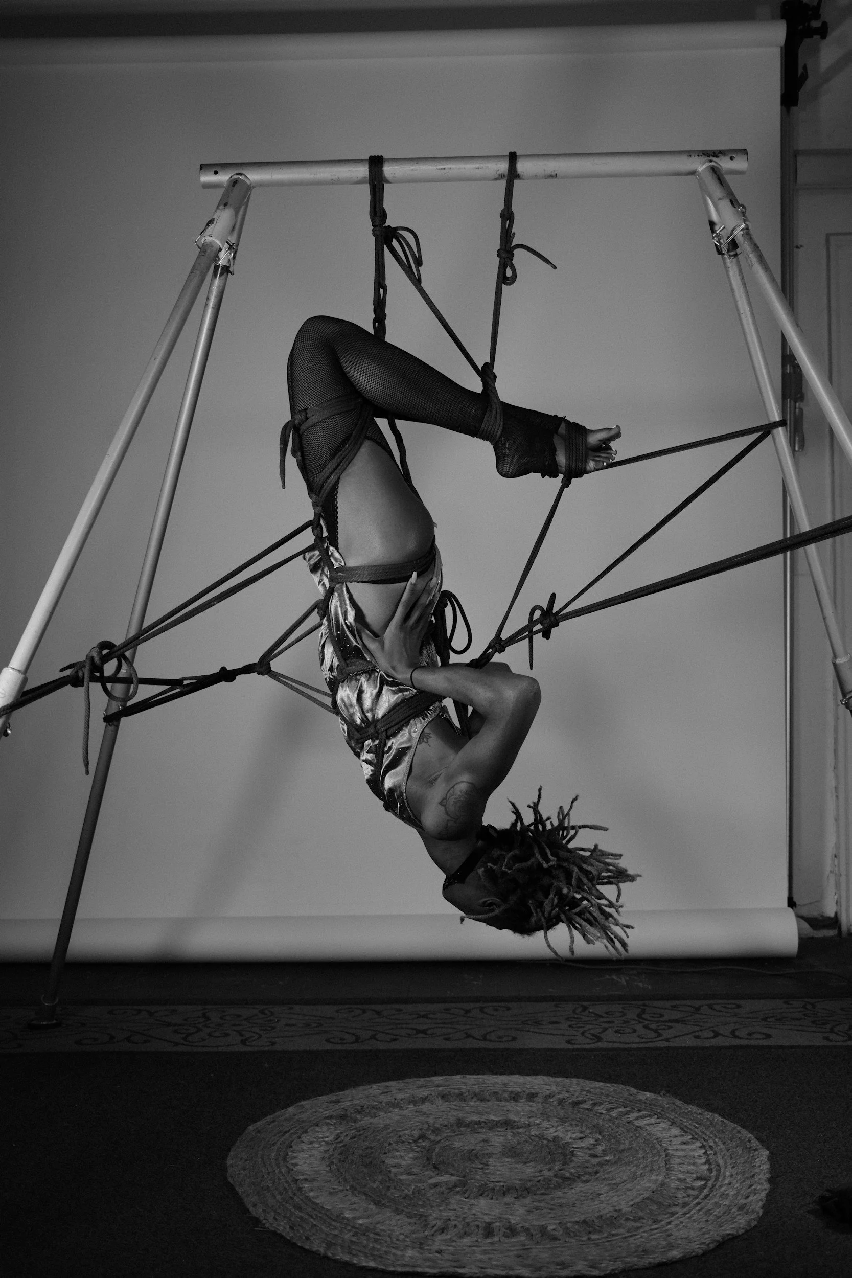 A woman performing an aerial yoga pose, hanging upside down from a metal structure with ropes, in a studio with a patterned rug on the floor.