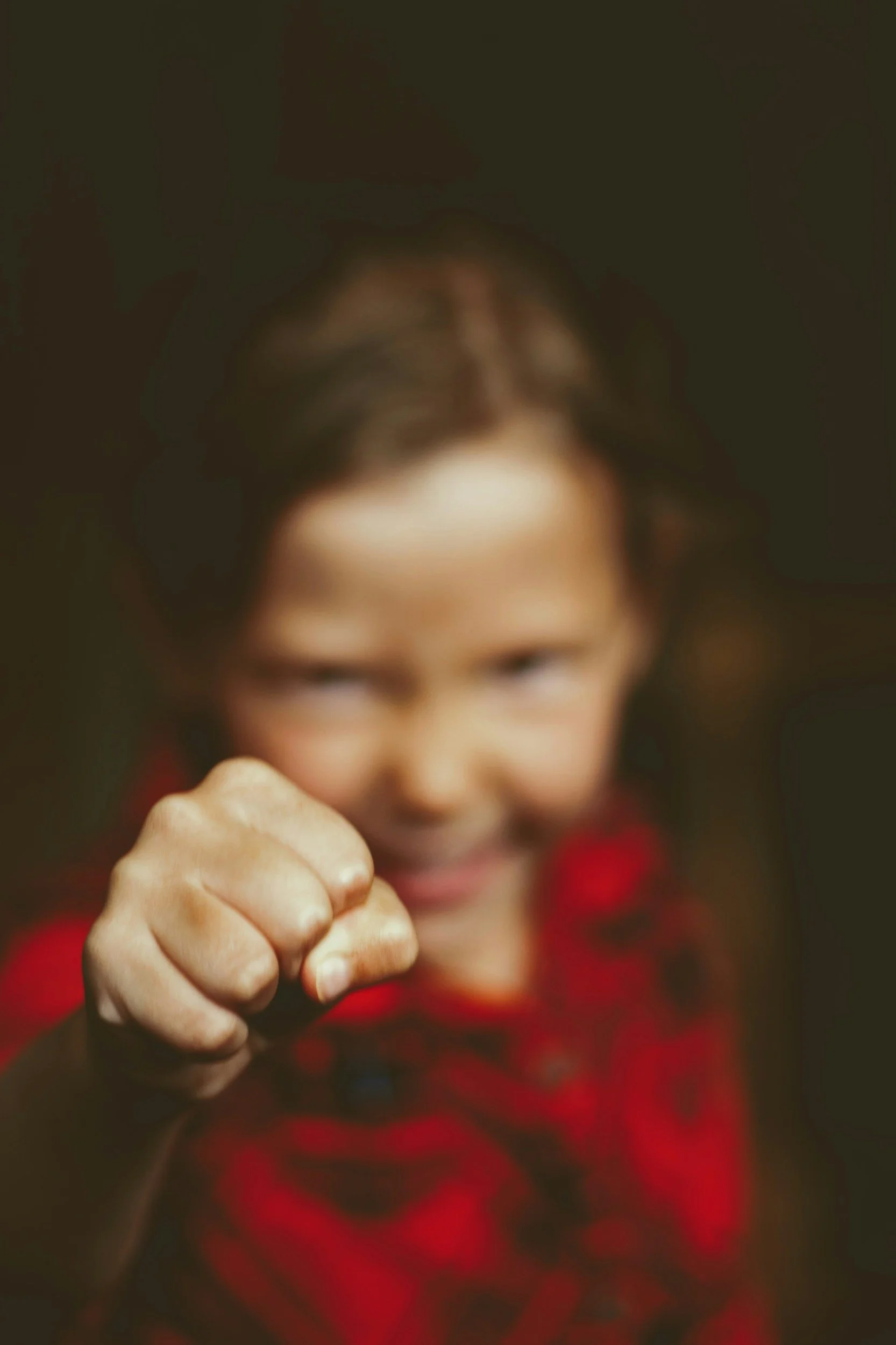 A young child with a focused expression, raising a clenched fist towards the camera, wearing a red shirt.