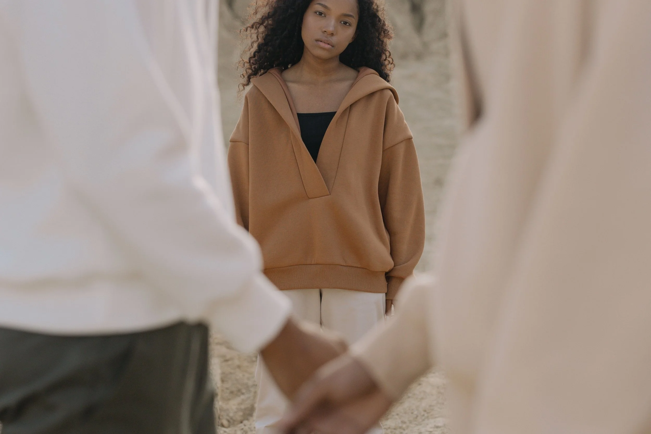A young woman with curly hair standing outdoors, wearing a tan hoodie and white pants, between two out-of-focus individuals.
