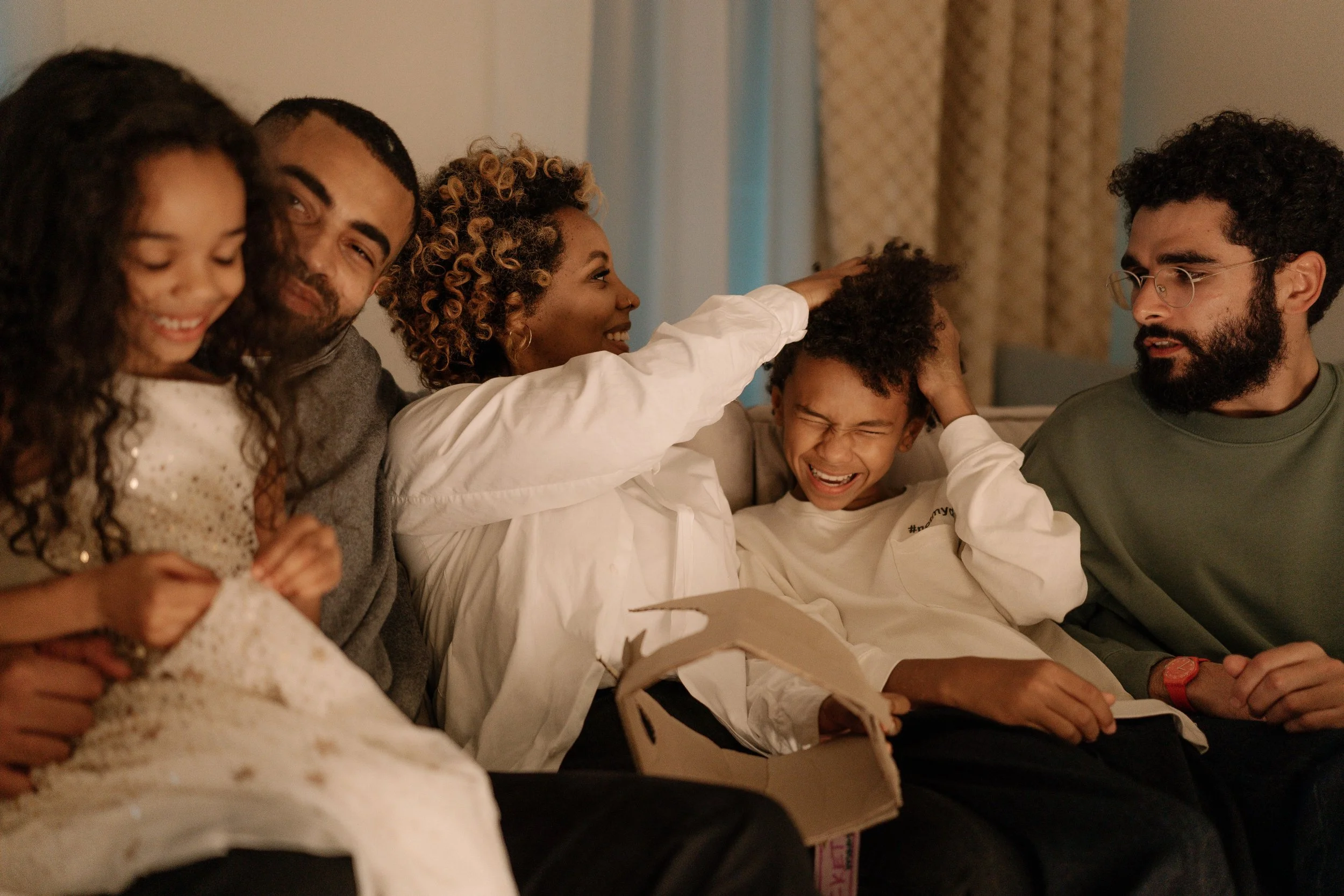 A diverse group of six people, including children and adults, sitting on a couch in a cozy living room, smiling and interacting with each other.